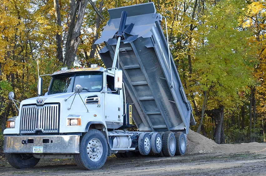 A large white dump truck unloading a pile of dirt at a construction site, surrounded by autumn trees.