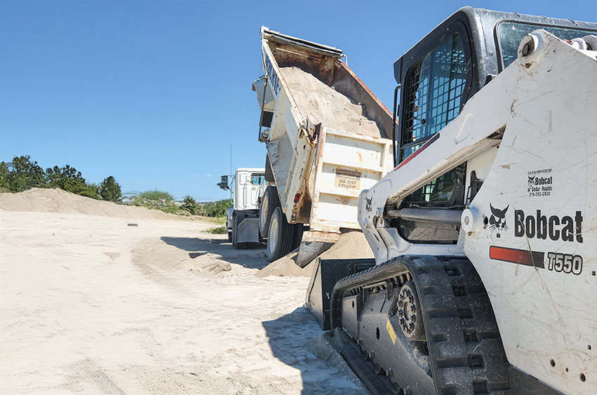 Bobcat and dump truck working together to haul sand at a job site.