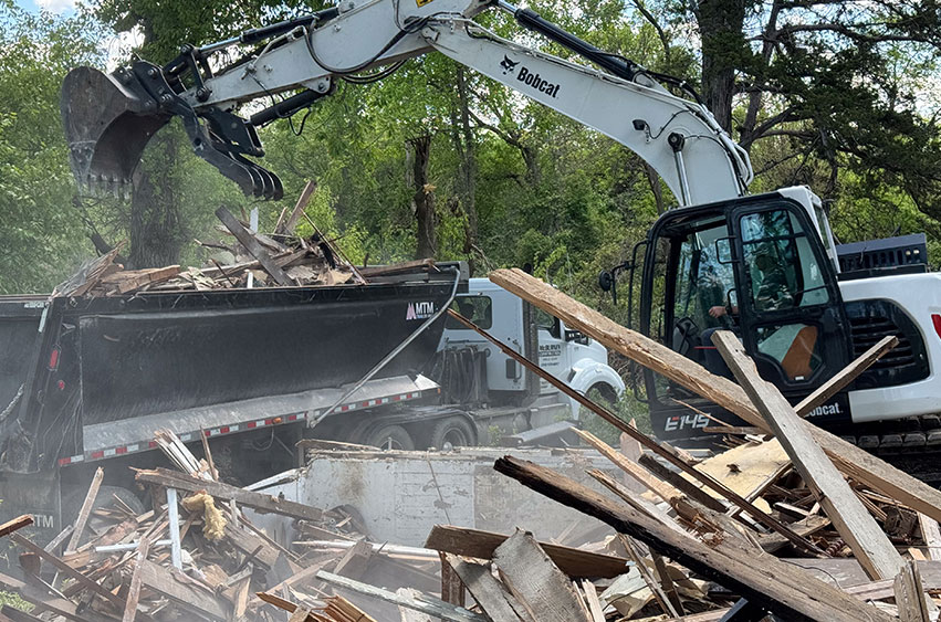 A Bobcat excavator loading wooden debris into a black dump trailer, clearing demolition waste from a site.