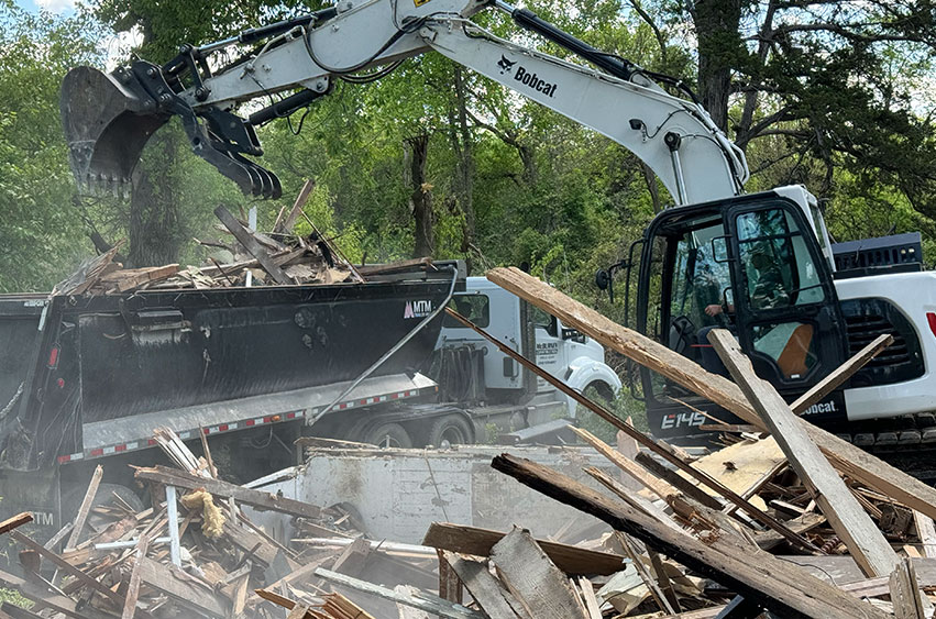 Excavator loading debris from a house demolition into a dump trailer for removal.