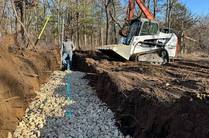 Worker measuring leach field trench lined with crushed stone while a skid loader moves material.